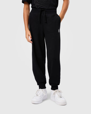 A person is shown from the shoulders down wearing black KIDS LEVY ESSENTIAL FLEECE SWEATPANTS by Psycho Bunny, featuring a tie at the waist and a small logo near the pocket. They are also wearing white sneakers and a black top. The background is plain and white.