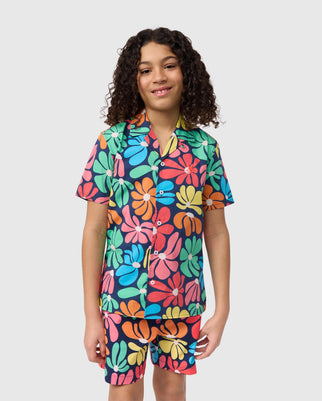 A young person with curly hair is smiling and wearing a vibrant Psycho Bunny KIDS MENTZ ALL OVER PRINT SHIRT - B0Q396B200 and matching shorts. The outfit, a regular fit featuring a colorful floral print in shades of blue, green, orange, pink, and yellow, makes for a perfect sun-soaked statement against the plain white background.