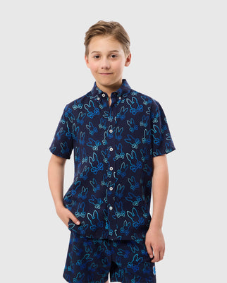 A young boy with short brown hair is wearing a navy blue Psycho Bunny KIDS SHELDON ALL OVER PRINT SHIRT (B0Q574C200) which features a playful bunny pattern in lighter blue tones. The Pima cotton fabric adds comfort as he stands with one hand in his pocket, smiling slightly, and looking directly at the camera against a plain light gray background.