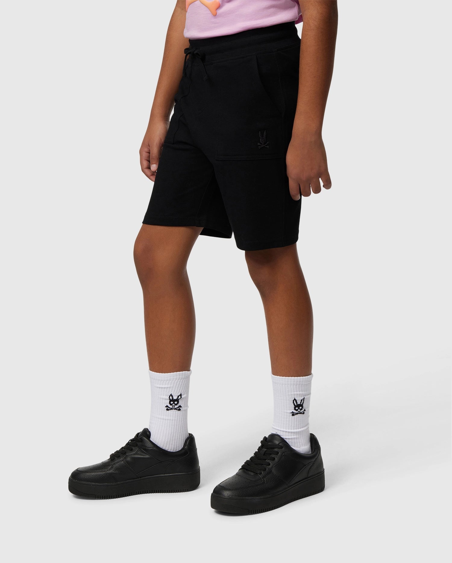 A person wearing a pink shirt made of Pima cotton piqué is shown from the neck down, dressed in black KIDS STANFORD LIGHT PIQUE SWEATSHORT - B0R344B200 by Psycho Bunny and black shoes. They are also wearing white calf-length socks with a small bunny logo in black. The background is plain white, highlighting this stylish wardrobe staple.