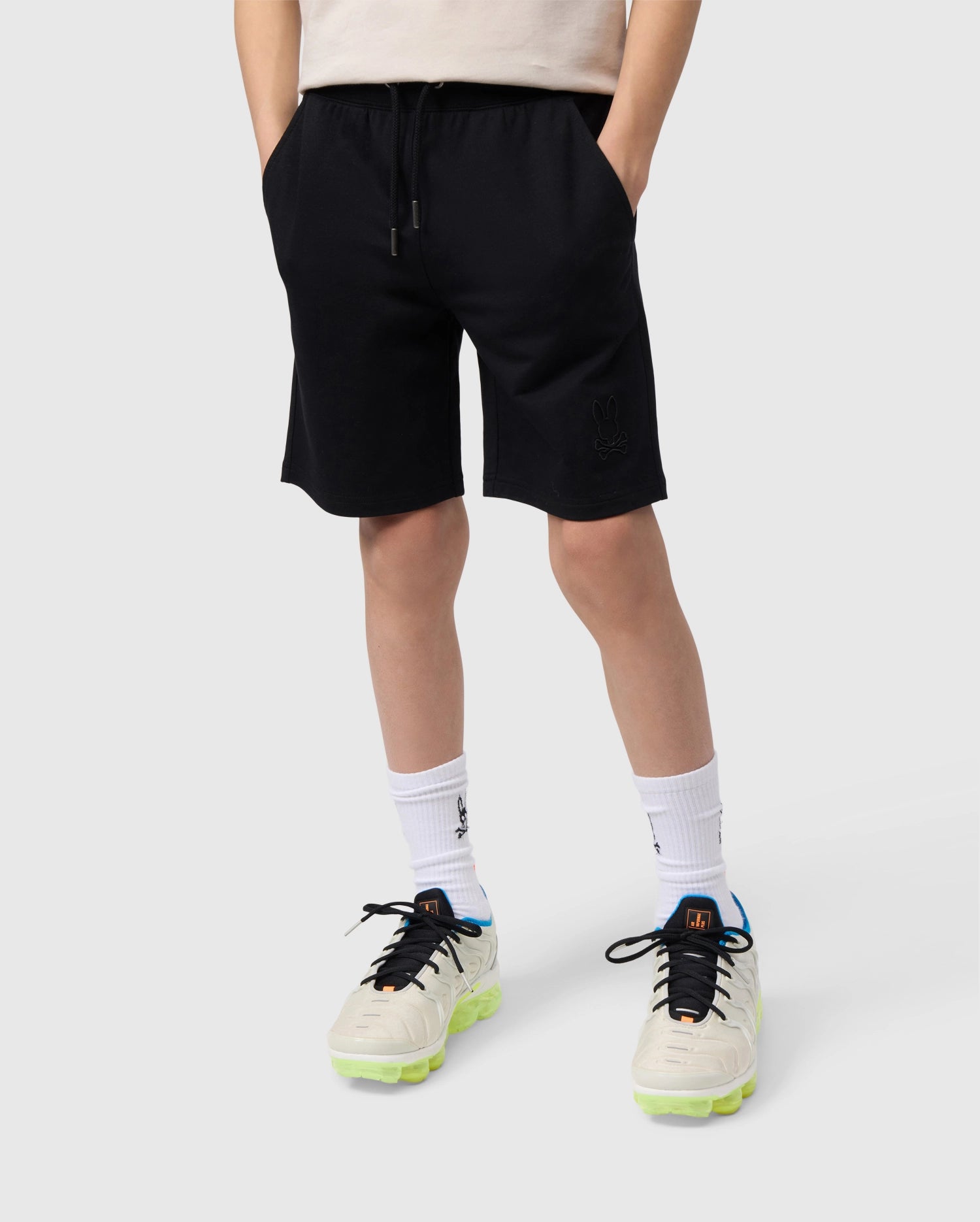 A person stands against a plain background, wearing black KIDS LIVINGSTON TERRY SWEATSHORT - B0R408B200 by Psycho Bunny, a beige top, white socks, and white sneakers with neon green soles. They have their hands in their pockets. The shorts, designed for comfort, feature a small emblem on the left leg.