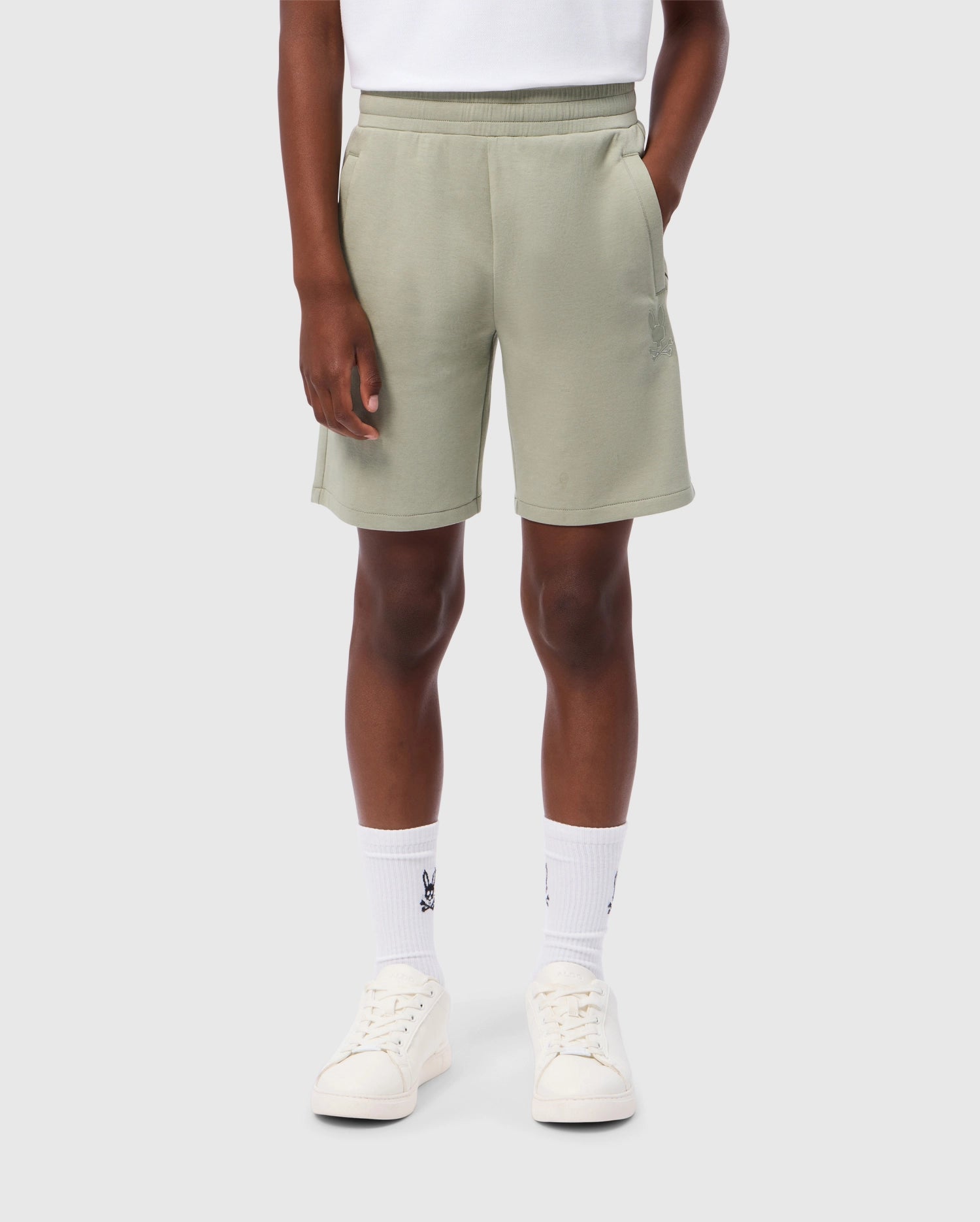 A person is wearing light green Psycho Bunny KIDS LYONS SWEATSHORT - B0R578C200 with hands in pockets, a white shirt, white calf-high socks with a dark emblem, and white sneakers. The background is plain and light gray.