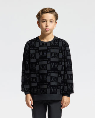 A young person poses against a simple backdrop, wearing a black KIDS ROMEO LIGHTWEIGHT SWEATSHIRT (B0S129E200) by Psycho Bunny. The sweatshirt features a bunny-patterned design with the brand's name "PSYCHO BUNNY" in a repeating pattern. Their short, light brown hair complements the look as they gaze directly at the camera.