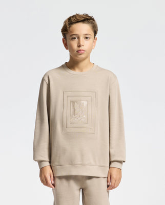 Against a plain white background, a young boy stands wearing the KIDS OAK CITY CREW NECK SWEATSHIRT by Psycho Bunny, paired with matching pants. This lightweight beige sweatshirt features a simple floral design in the center that complements his short light brown hair and neutral expression.