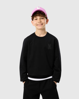 A smiling boy wearing the Psycho Bunny KIDS HOUSTON FRENCH TERRY CREWNECK - B0S462C200, crafted from premium cotton fabric, paired with black pants. His curly brown hair peeks from under a pink cap. With hands in pockets, he stands against a plain gray background, showcasing style and comfort in every detail.