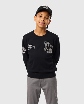 A person wearing the Psycho Bunny KIDS ZACK VARSITY SWEATSHIRT - B0S880D200, in black with chenille embroidery, along with a matching black cap, stands against a plain background. The sweatshirt showcases writing on the chest and left sleeve, as well as a vintage-inspired logo. The person is smiling with their hands behind their back.