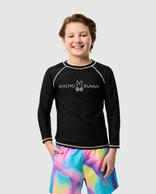 A child smiles wearing the black Psycho Bunny kids' long sleeve rash guard (B0T557C200) with a bunny logo, paired with colorful tie-dye shorts in pink, blue, and yellow. The background is plain white.