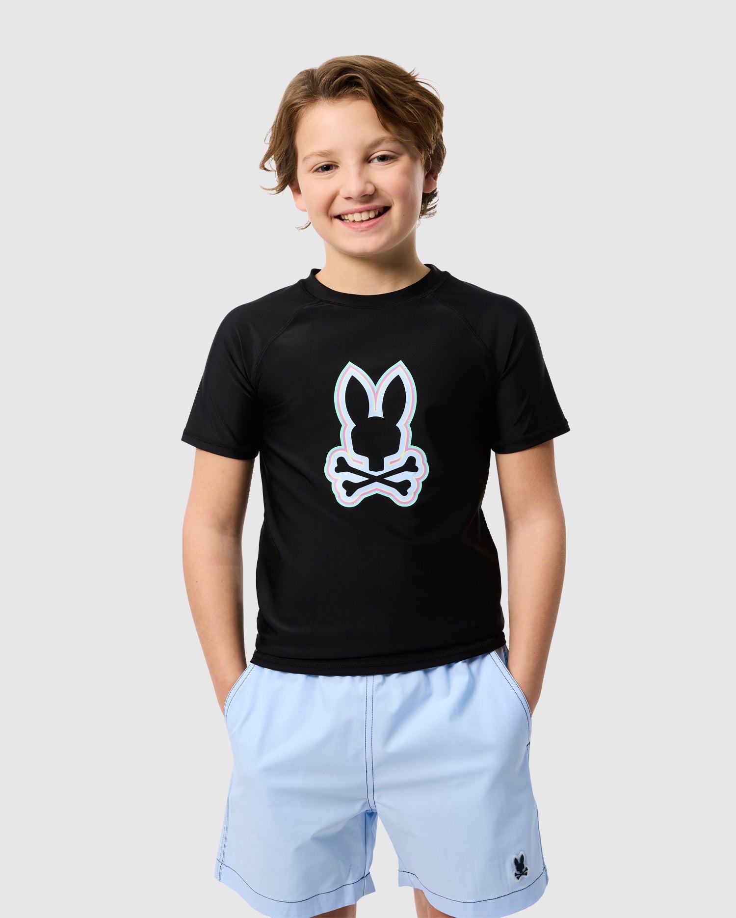 A joyful young boy wearing a Psycho Bunny KIDS SAN LEON ANTI-UV RASHGUARD - B0T645C200 with a white rabbit logo and light blue shorts, standing against a plain white background and smiling at the camera.