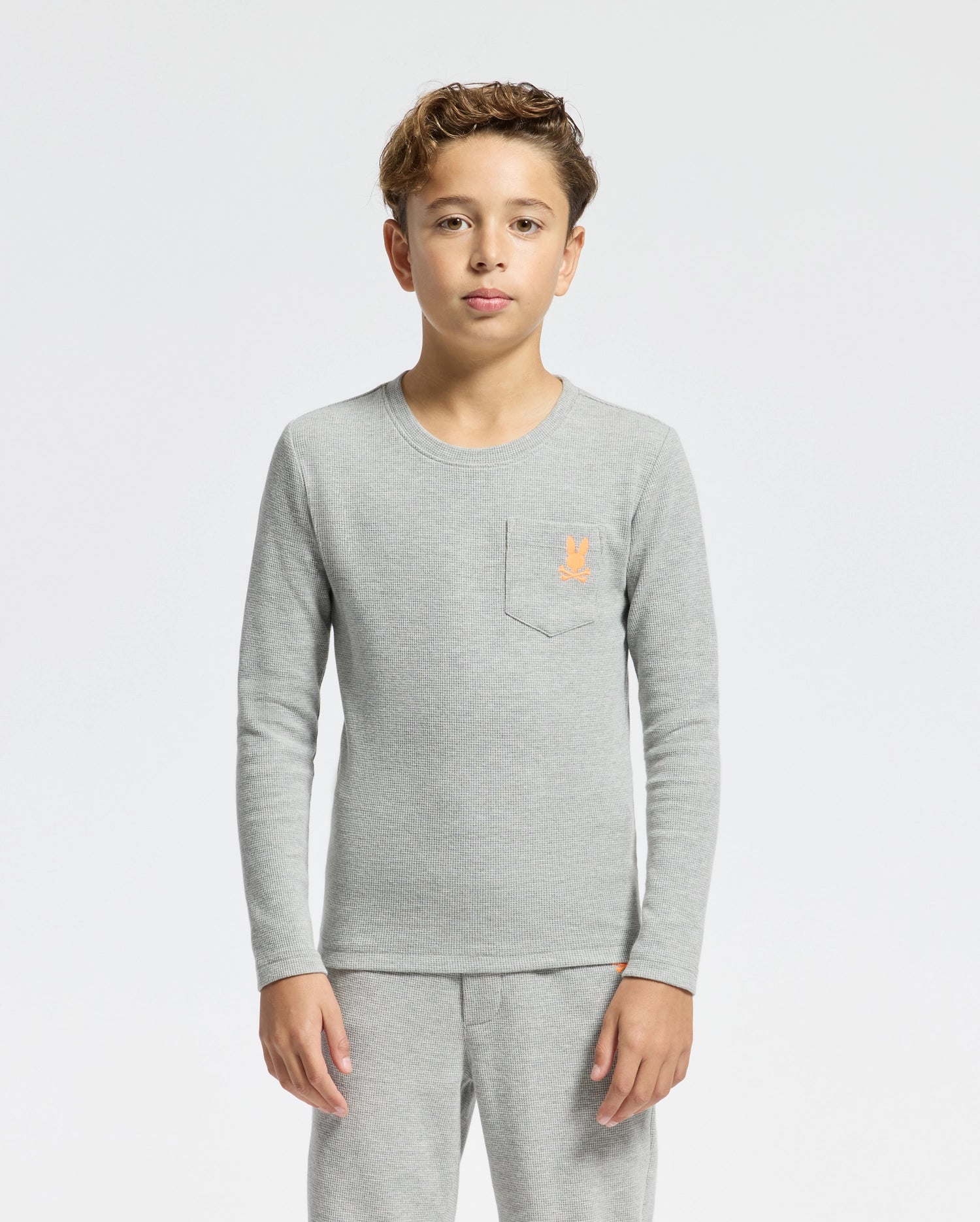 A young boy with short curly hair is wearing a light gray KIDS VAL WAFFLE LONG SLEEVE TEE - B0T672E200 by Psycho Bunny and pants made of soft Pima Cotton. An embroidered orange logo decorates the chest pocket, perfectly complementing the plain light gray background.