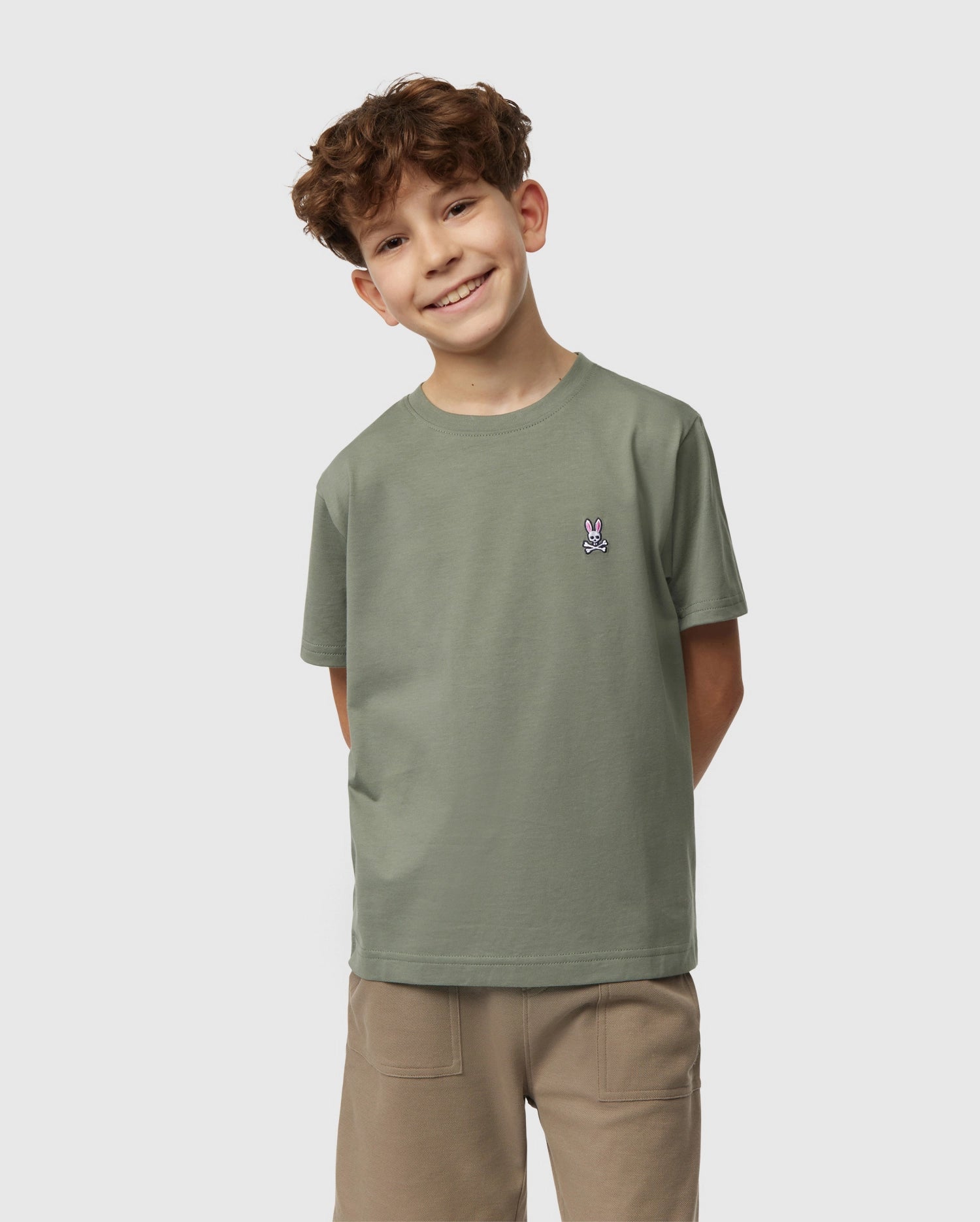 A smiling child with curly brown hair stands against a plain white background. They are wearing a light green Psycho Bunny Kids Classic Crew Neck Tee (B0U014B200) made from 100% Pima cotton, featuring a small rabbit logo on the left side, paired with beige shorts—the perfect back-to-school outfit. Their hands are behind their back.