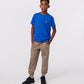 A young boy stands confidently with his hands in his pants pockets, wearing a bright blue KIDS CLASSIC CREW NECK TEE by Psycho Bunny, crafted from soft Pima cotton. Beige jogger pants and black sneakers complete his look against a plain white background.