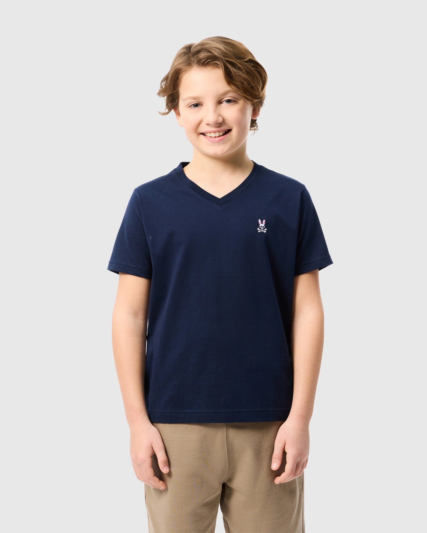 A smiling young person with short, light brown hair is wearing the KIDS CLASSIC V NECK TEE - B0U100CRPC from Psycho Bunny, a navy T-shirt made of soft Pima cotton adorned with a small emblem on the chest. They have paired it with khaki pants and are standing against a plain light gray background, perfect for the back-to-school season.