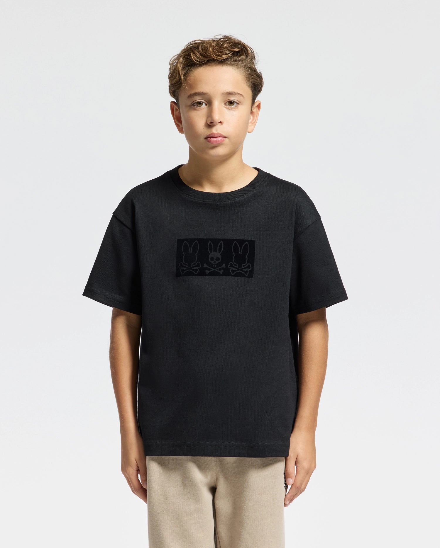 A young boy with short, curly hair stands against a plain background. He is wearing the KIDS ROMEO HEAVY WEIGHT RELAXED FIT TEE - B0U110E200 from Psycho Bunny, crafted from soft pima cotton with a subtle print of three bunnies. His beige pants complement the heavyweight T-shirt, and his expression remains neutral.