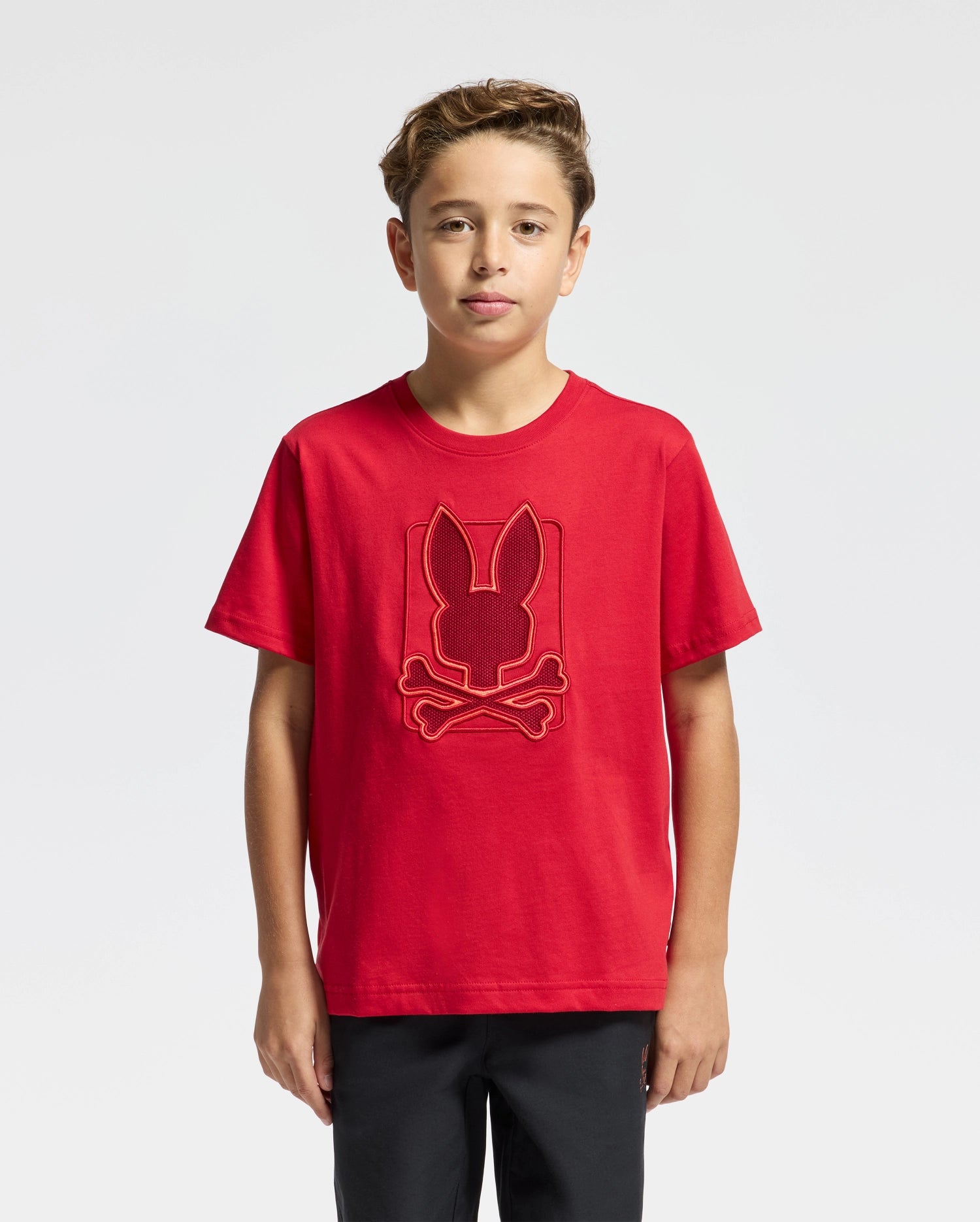 A young person is wearing the KIDS PIERCE GRAPHIC TEE - B0U115E200 from Psycho Bunny, crafted from soft pima cotton. The red shirt features a bunny embroidery with a crossbones design. They stand against a plain white background, have short light brown hair, and are also wearing dark pants.