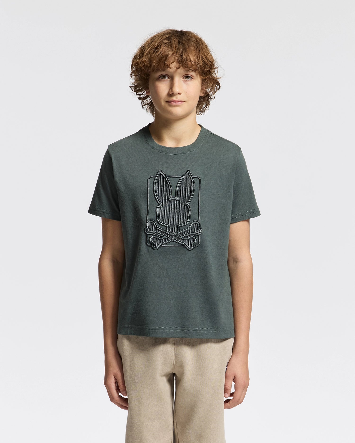 A person with curly brown hair is standing against a plain white background, wearing a dark green KIDS PIERCE GRAPHIC TEE - B0U115E200 by Psycho Bunny paired with beige pants.