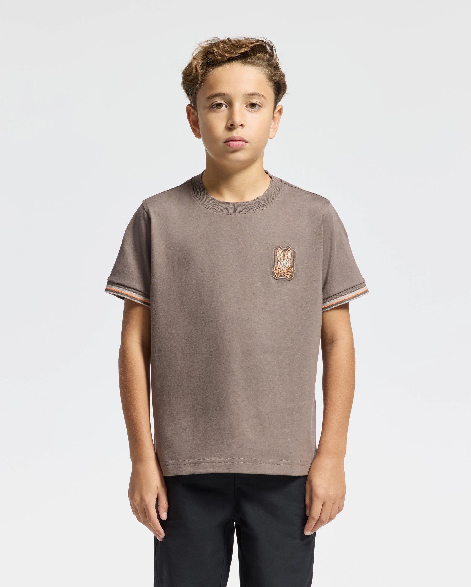 A young boy with short hair is wearing a KIDS PIERCE FASHION TEE - B0U140E200, crafted from soft Pima cotton by Psycho Bunny, featuring a small embroidered patch of a bunny on the chest. The background is plain white, and he stands with his arms at his sides, looking confidently at the camera.