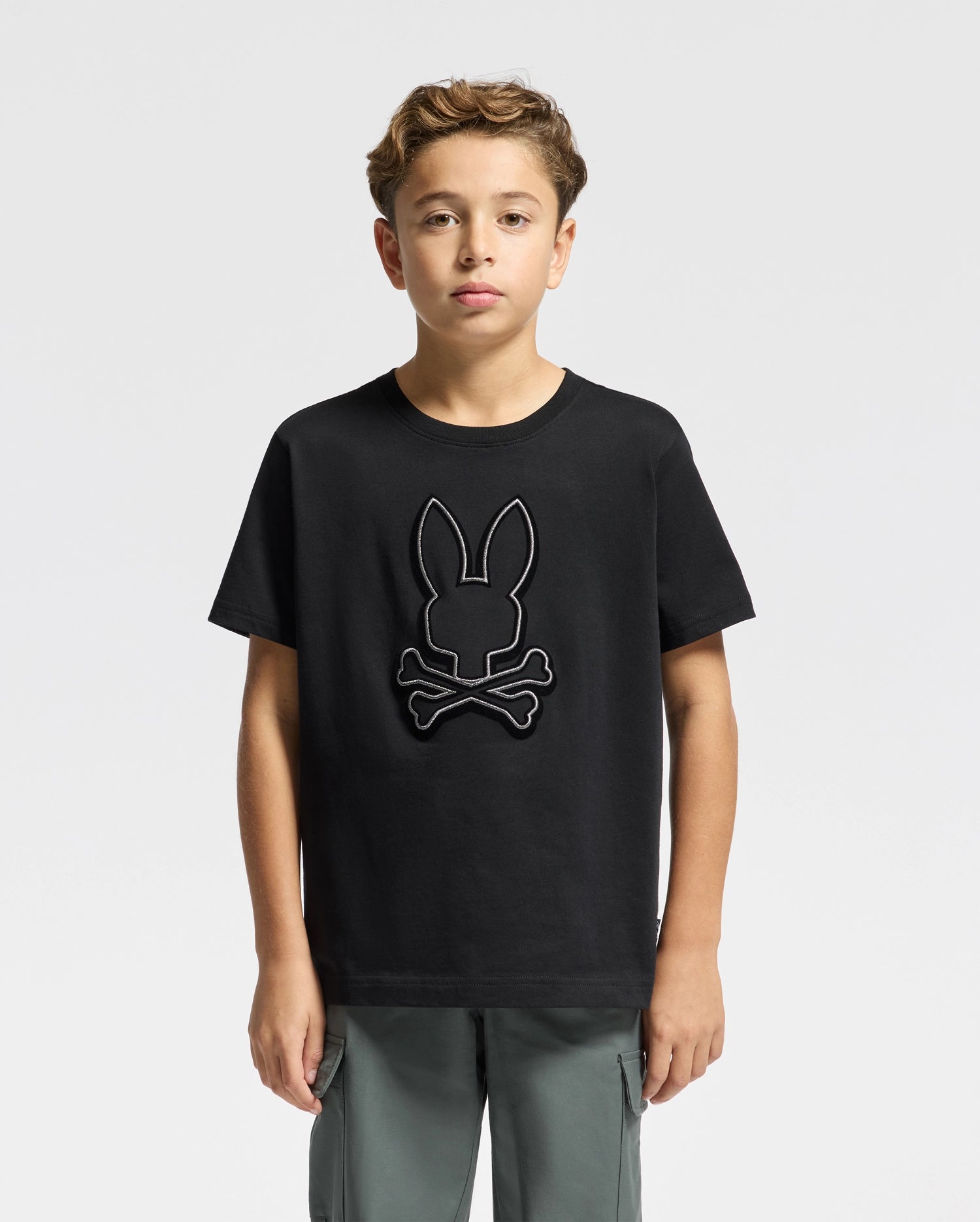 A young boy stands facing forward, wearing the KIDS ALEXANDER GRAPHIC TEE - B0U163E200 by Psycho Bunny, featuring a bunny and crossbones design. Made from premium Pima Cotton, the black T-shirt is ideally matched with gray pants against a plain white background.