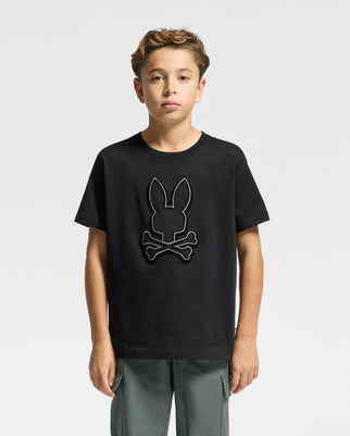 A young boy stands facing forward, wearing the KIDS ALEXANDER GRAPHIC TEE - B0U163E200 by Psycho Bunny, featuring a bunny and crossbones design. Made from premium Pima Cotton, the black T-shirt is ideally matched with gray pants against a plain white background.