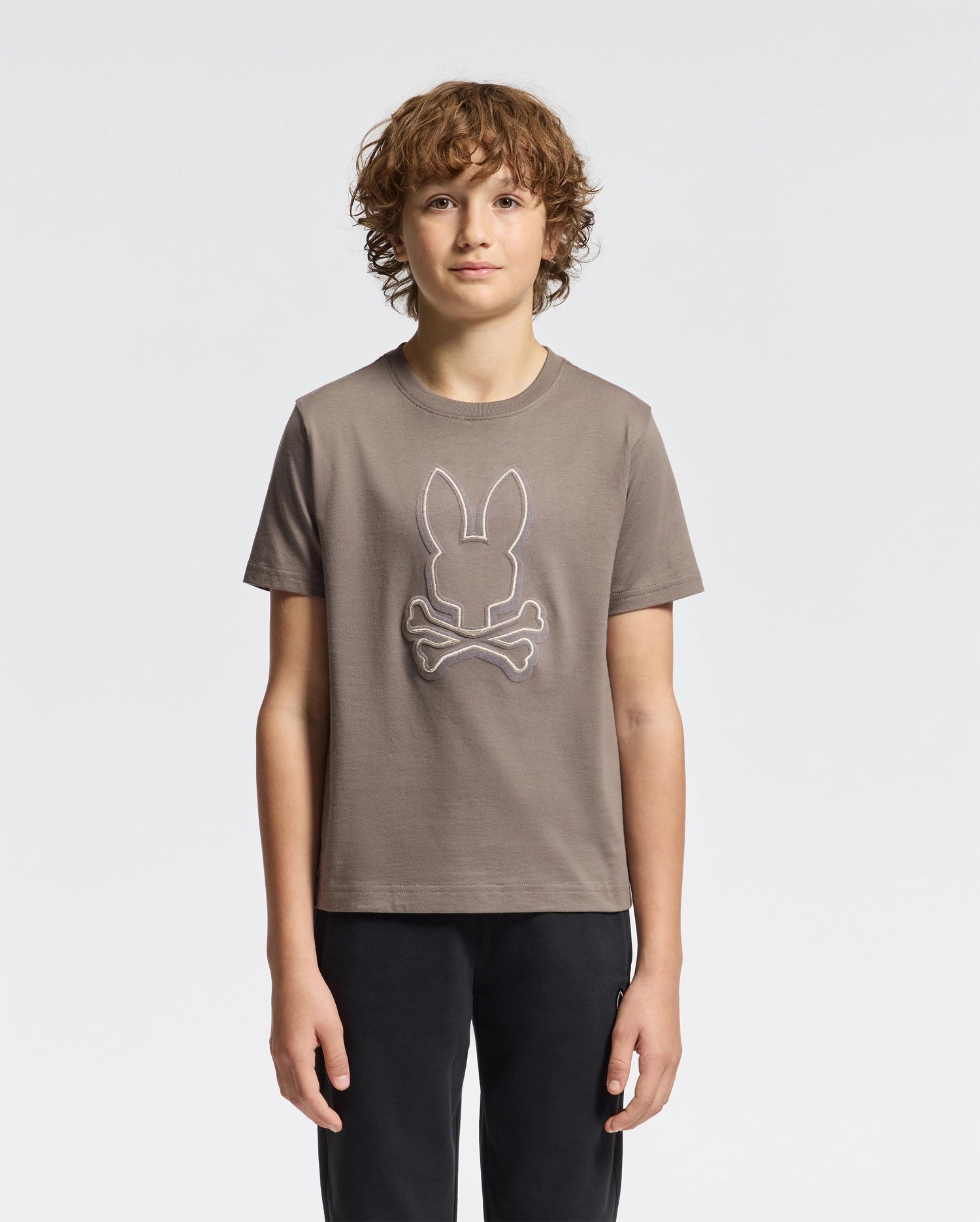 A young person with curly brown hair stands against a plain white background, wearing the Psycho Bunny KIDS ALEXANDER GRAPHIC TEE - B0U163E200 in taupe, which features a white outline of a rabbit head above crossed bones, paired with black pants.