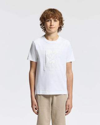 A young person with curly hair stands against a light gray background wearing a plain white KIDS DOTSERO PRINTED GRAPHIC TEE by Psycho Bunny, crafted from soft Peruvian Pima Cotton, and complemented by beige pants. The T-shirt showcases a subtle embossed floral design.