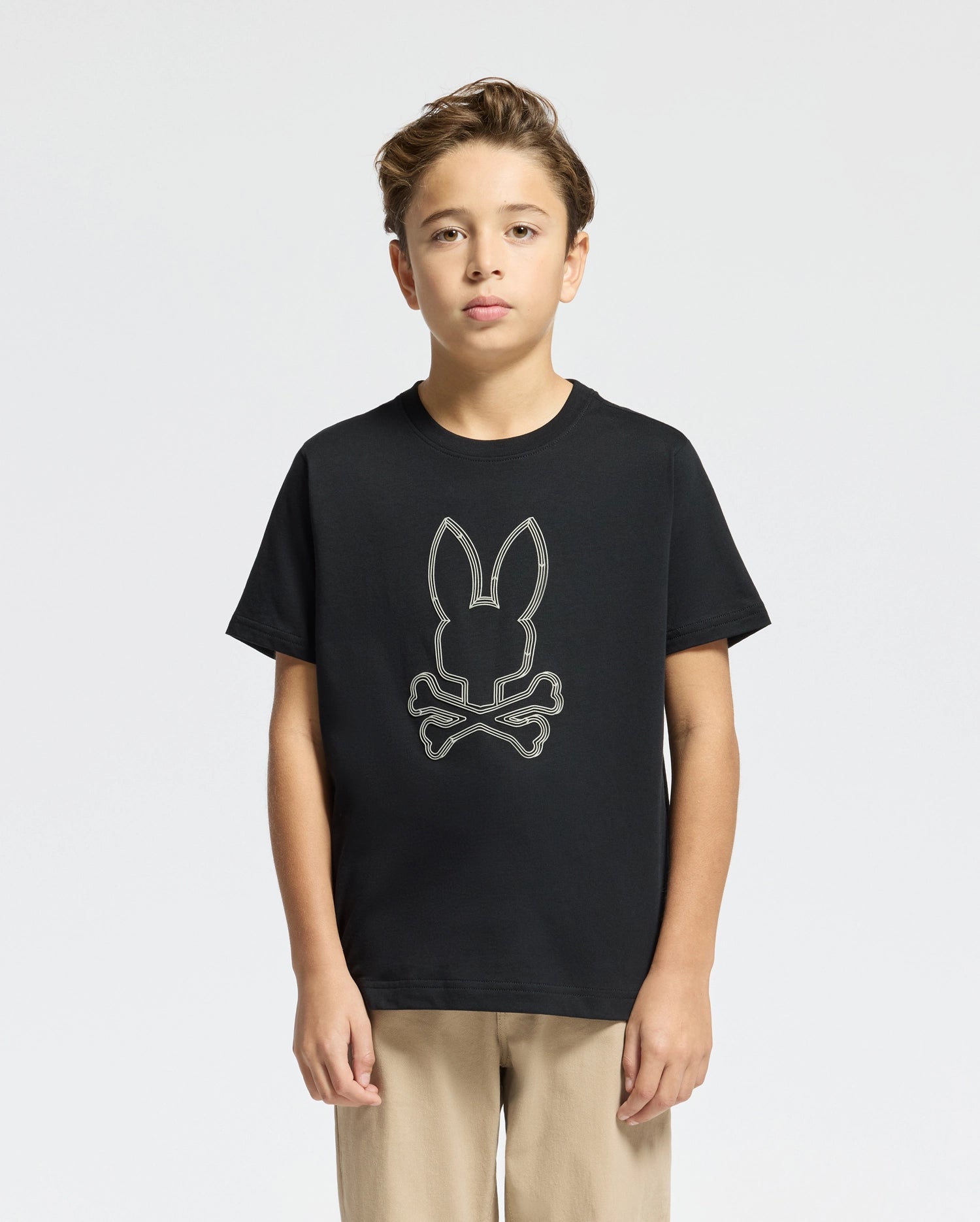 A young boy stands wearing a Psycho Bunny KIDS MONARC FRONT BUNNY GRAPHIC TEE - B0U211E200, crafted from soft Pima cotton and featuring a skull and crossbones design. He has short, light brown hair and is dressed in beige pants against a plain white background.