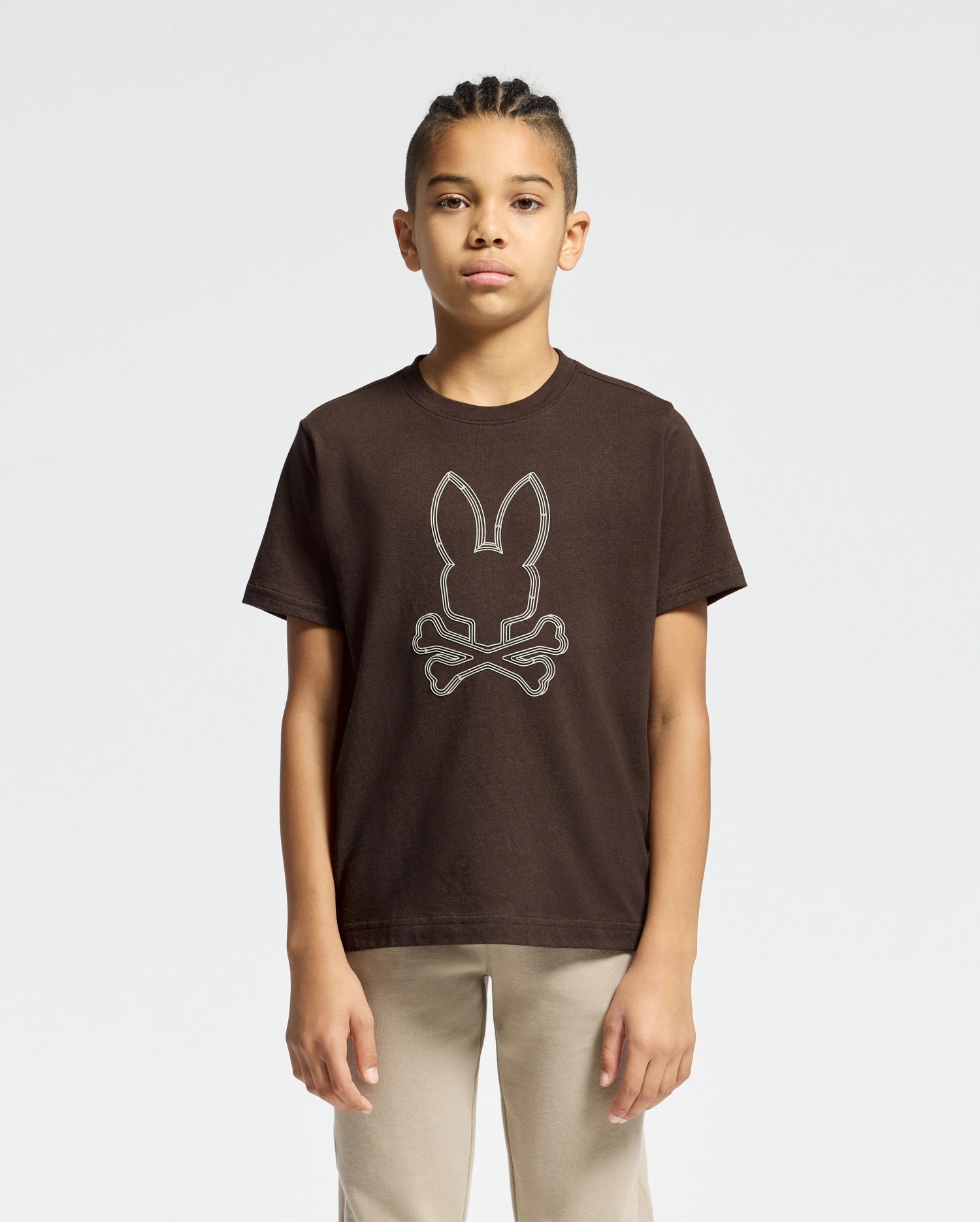 A young person with braided hair stands against a plain background, wearing a brown KIDS MONARC FRONT BUNNY GRAPHIC TEE - B0U211E200 by Psycho Bunny, featuring a rabbit head above crossed bones. Made from soft Pima Cotton, they maintain a neutral expression and are dressed in beige pants.