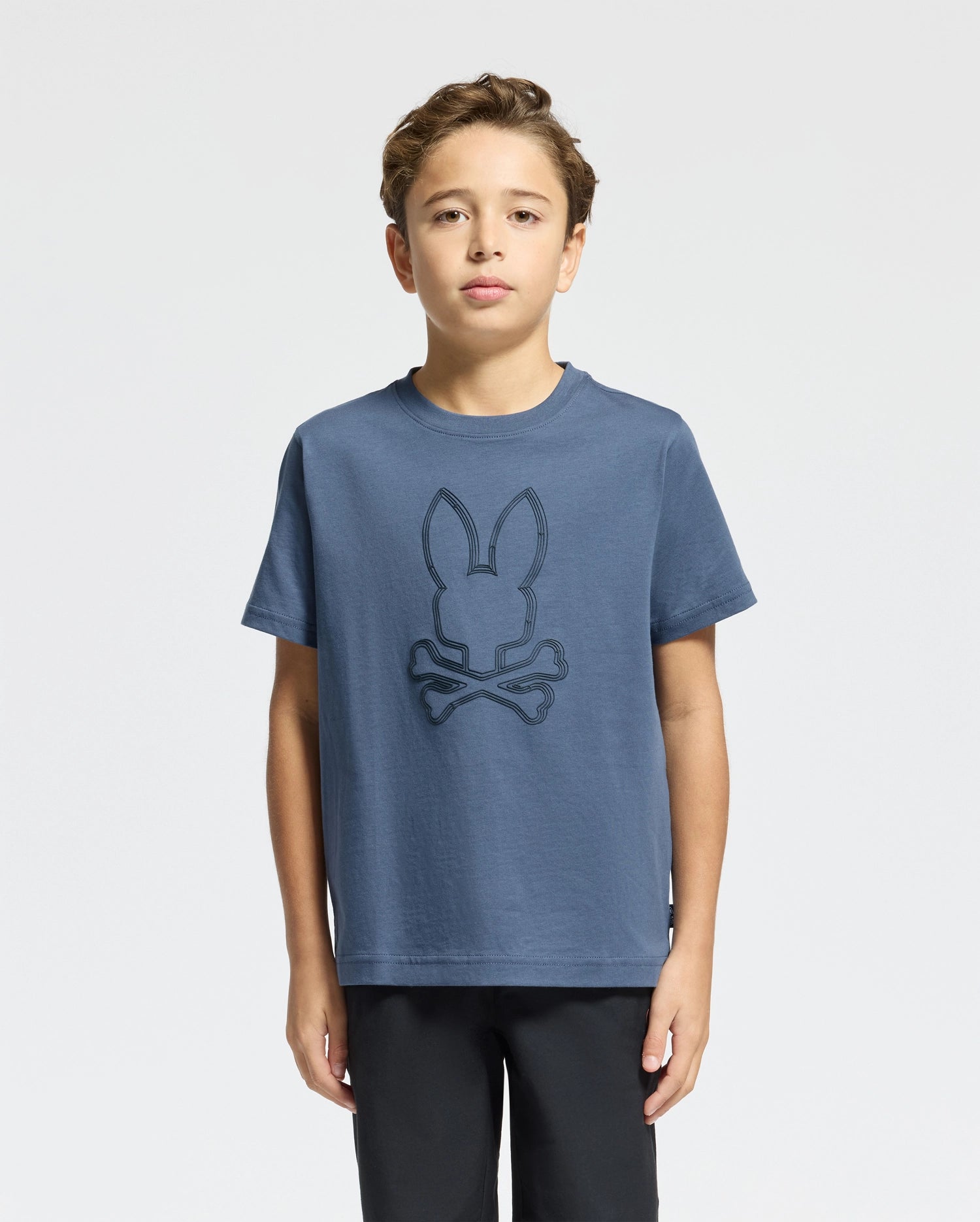 A young boy is wearing the KIDS MONARC FRONT BUNNY GRAPHIC TEE - B0U211E200 by Psycho Bunny, crafted from soft Pima cotton. The tee showcases a design with bunny ears and crossbones. His short, wavy hair frames his face as he gazes directly at the camera, set against a simple light background.