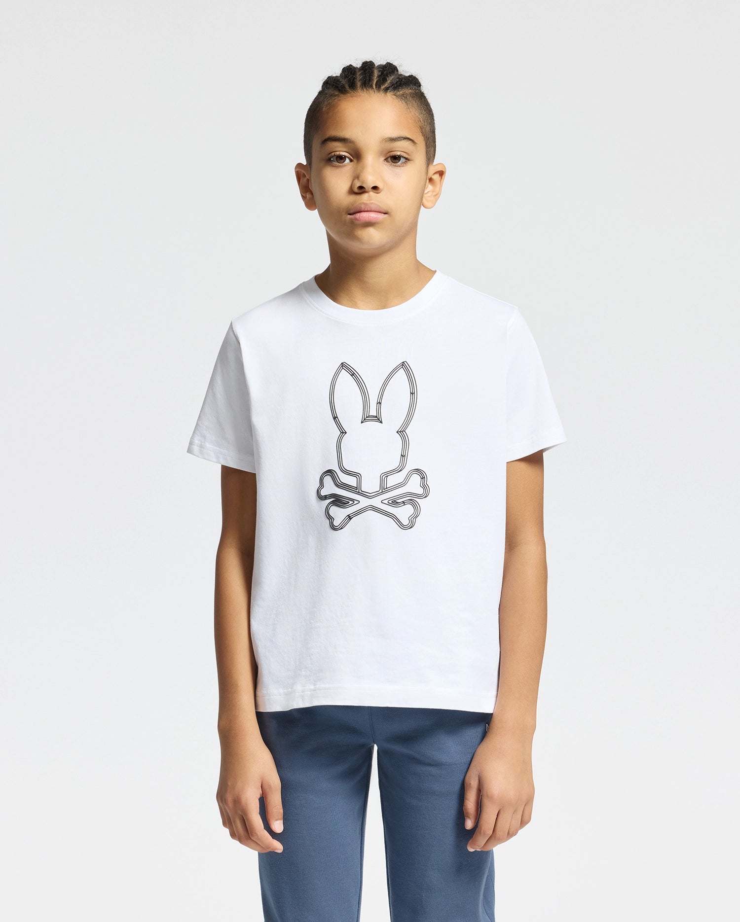 A young individual with braided hair is wearing the KIDS MONARC FRONT BUNNY GRAPHIC TEE - B0U211E200 by Psycho Bunny, crafted from soft Pima Cotton and showcasing a rabbit head logo above crossed bones. They are also sporting blue pants and are positioned against a plain white background.