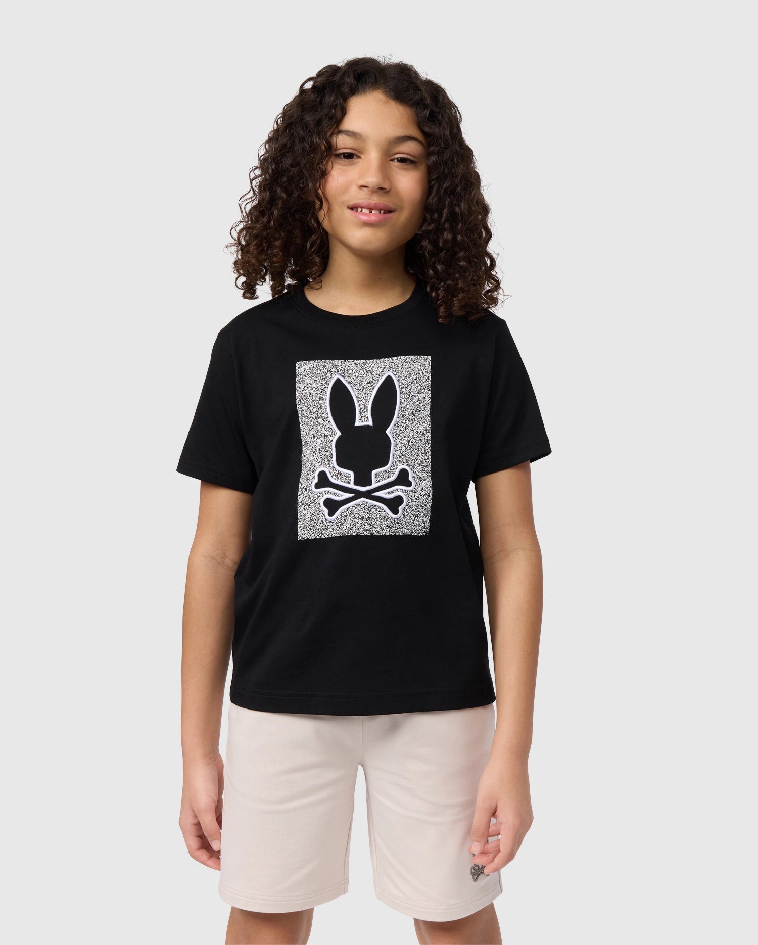 A smiling child with curly hair is wearing a black KIDS LIVINGSTON GRAPHIC TEE - B0U247B2TS by Psycho Bunny featuring a bunny skull and crossbones design. They are also wearing light-colored shorts and standing against a plain light gray background.
