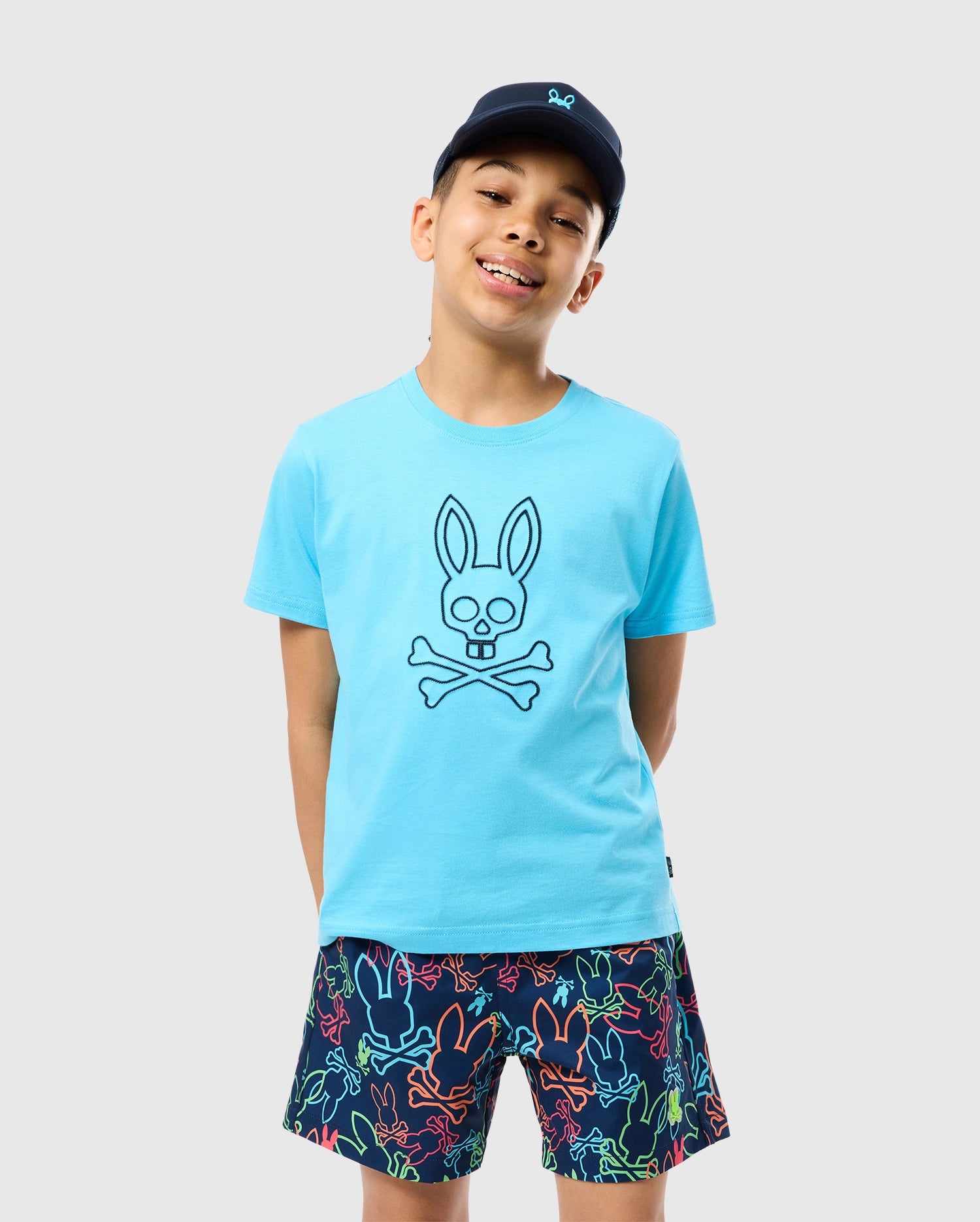 A young boy wearing a light blue Psycho Bunny KIDS SHELDON GRAPHIC TEE - B0U569C200 featuring a skull and crossbones design with bunny ears stands against a plain background. He sports a navy blue cap and colorful shorts with a bunny pattern in various neon colors, smiling with his hands behind his back.