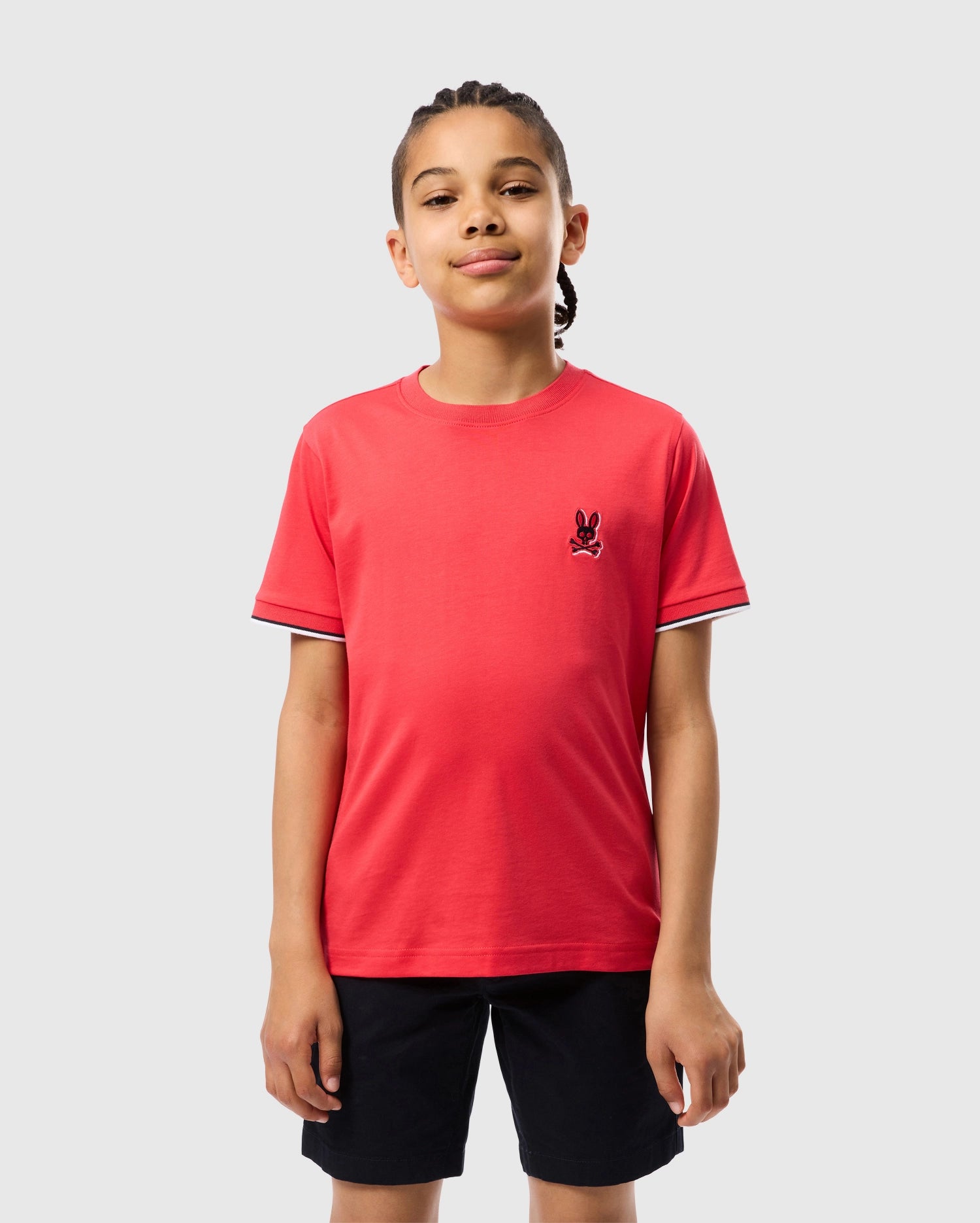 A young boy with braided hair stands against a plain background, wearing a bright red KIDS KAYDEN FASHION TEE - B0U577C200 by Psycho Bunny, crafted from Pima cotton. The shirt features an embroidered chest Bunny logo on the left side of the chest and is paired with black shorts. He is smiling slightly with his hands relaxed at his sides.