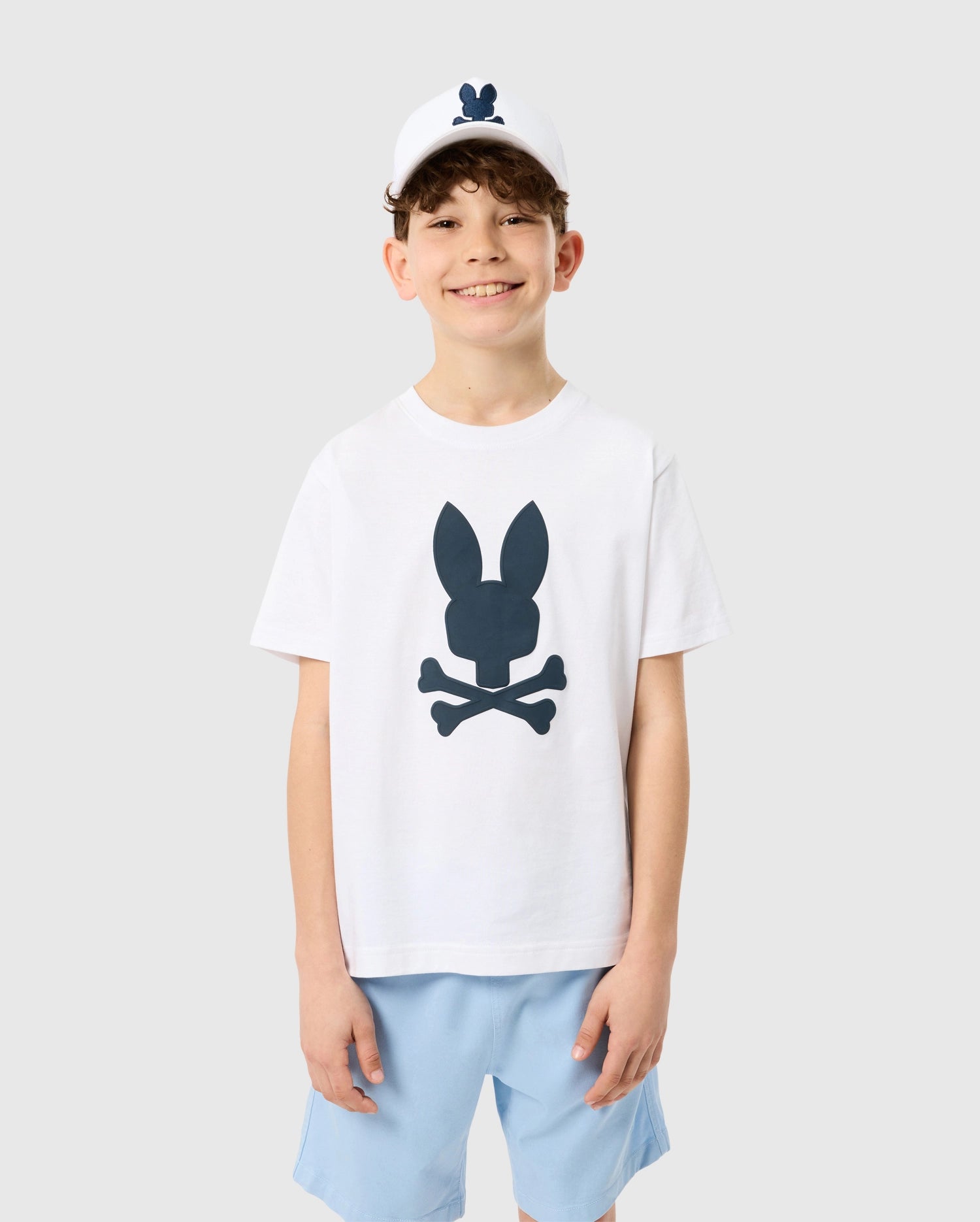 A smiling boy wearing a white KIDS HOUSTON GRAPHIC TEE by Psycho Bunny, featuring a black bunny and crossbones design, paired with light blue shorts and a white cap with a bunny logo, standing against a grey background.