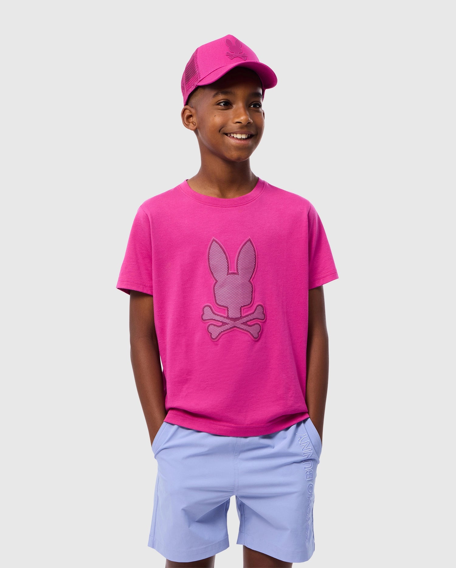 A young boy stands against a plain white background, hands in his pockets, wearing a cheerful smile. He sports the Psycho Bunny KIDS RIVIERA GRAPHIC TEE - B0U639C200, a bright pink t-shirt crafted from soft Peruvian Pima cotton with a bunny and crossbones design. His outfit is completed with a matching pink cap and light blue shorts.