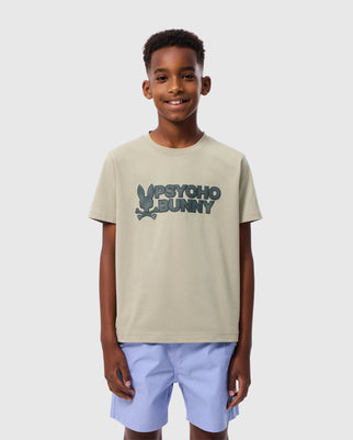 A young boy stands smiling, wearing a light beige, Pima cotton KIDS MELVIN GRAPHIC TEE - B0U663C200. The t-shirt features the "Psycho Bunny" brand name and bunny logo in dark green lettering, with an HD-silicone print wordmark graphic. He pairs it with light blue shorts against a plain white background.