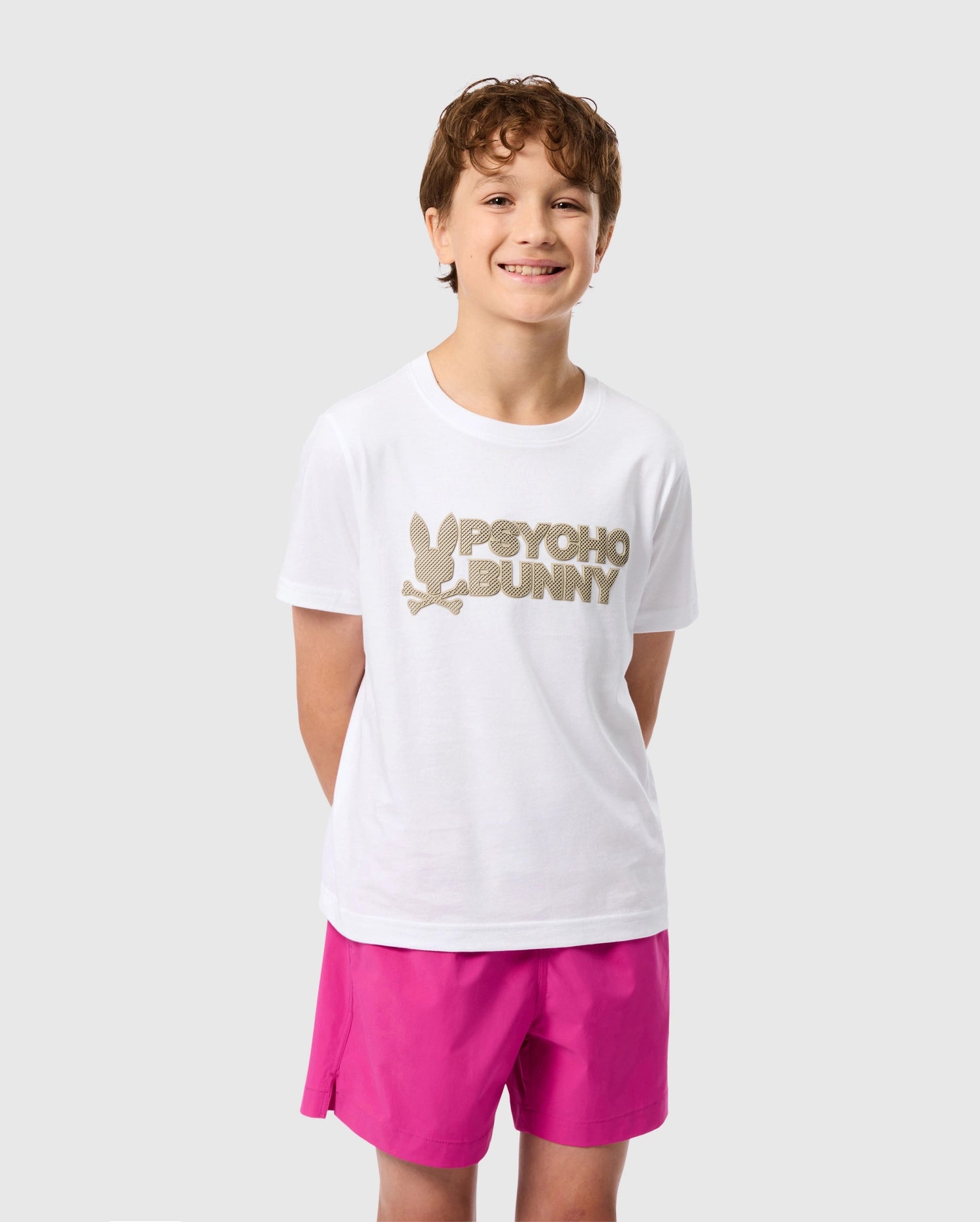 A smiling young boy stands with his hands behind his back against a plain background. He is wearing a white KIDS MELVIN GRAPHIC TEE - B0U663C200 by Psycho Bunny, featuring a gold 