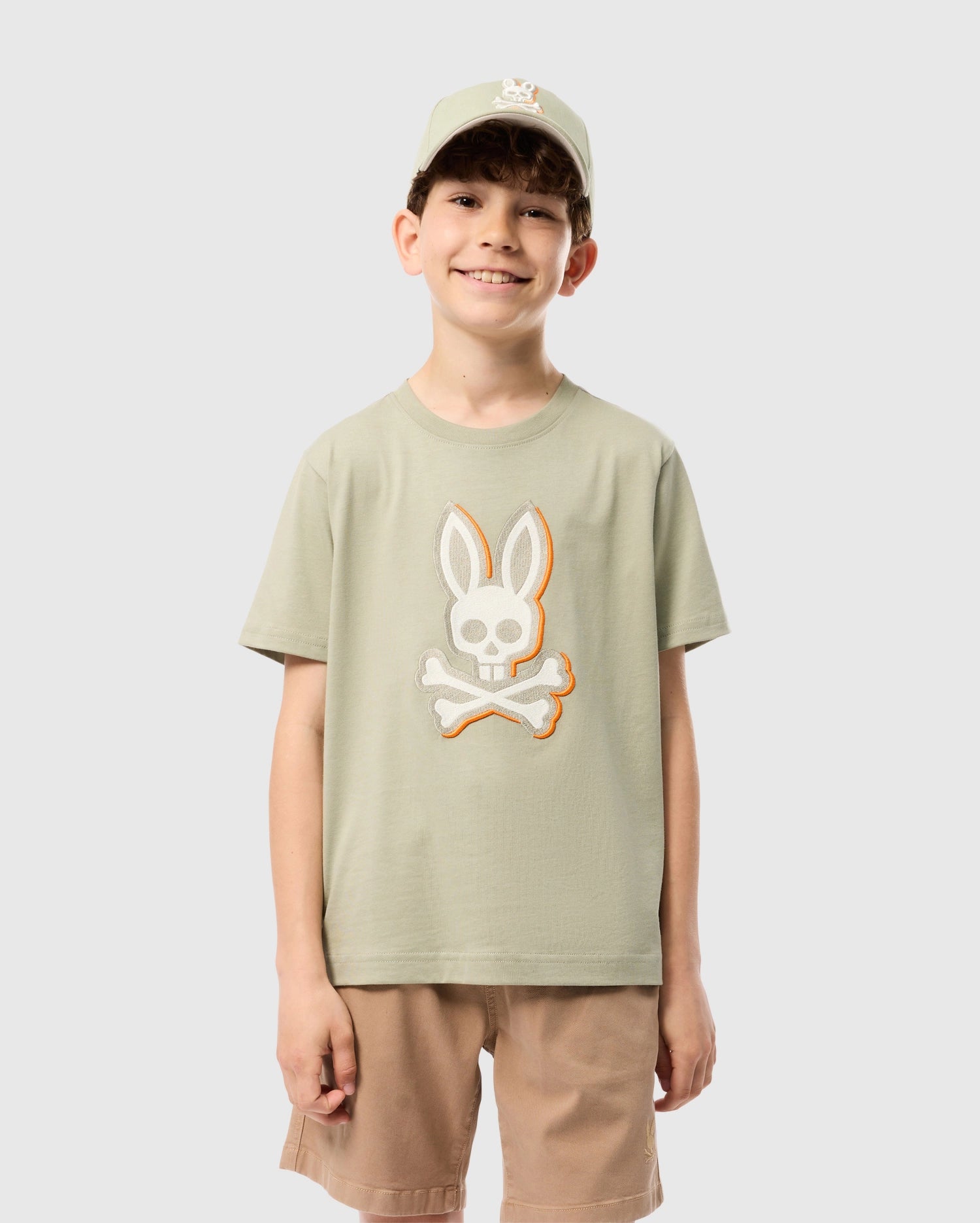 A young boy is smiling at the camera. He is wearing a Psycho Bunny KIDS KAYDEN GRAPHIC TEE - B0U676C200 in sage green, adorned with a bunny skull and crossbones on the front. He pairs it with beige shorts and a matching sage green baseball cap, also featuring the bunny embroidered logo.