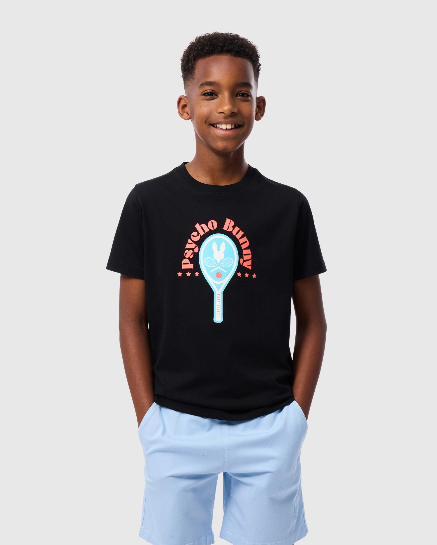 A smiling boy stands against a gray background, wearing the KIDS COURTSIDE GRAPHIC TEE by Psycho Bunny, featuring a blue tennis racket and 