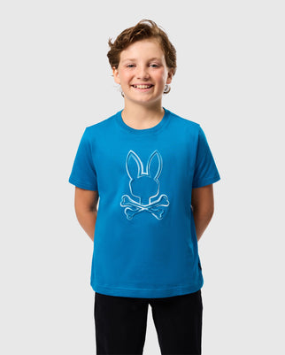 A smiling young boy with wavy hair wears the Psycho Bunny KIDS VINCENT GRAPHIC TEE - B0U713D200, a blue regular fit t-shirt made from soft Pima cotton that features a rabbit head above crossed bones. His hands are behind his back as he stands against a plain light gray background.