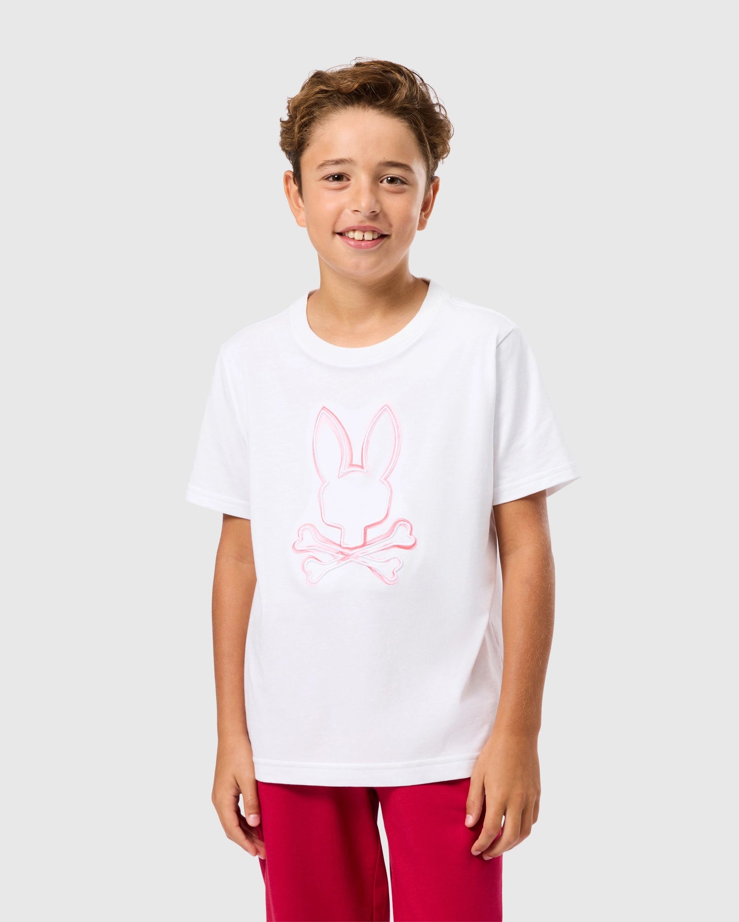 A smiling boy with short curly hair is standing against a plain background. He is wearing a Psycho Bunny KIDS VINCENT GRAPHIC TEE - B0U713D200, which features a pink outline of a rabbit above crossbones on the front, and red pants. His outfit exudes an unfadable style that's both playful and trendy.