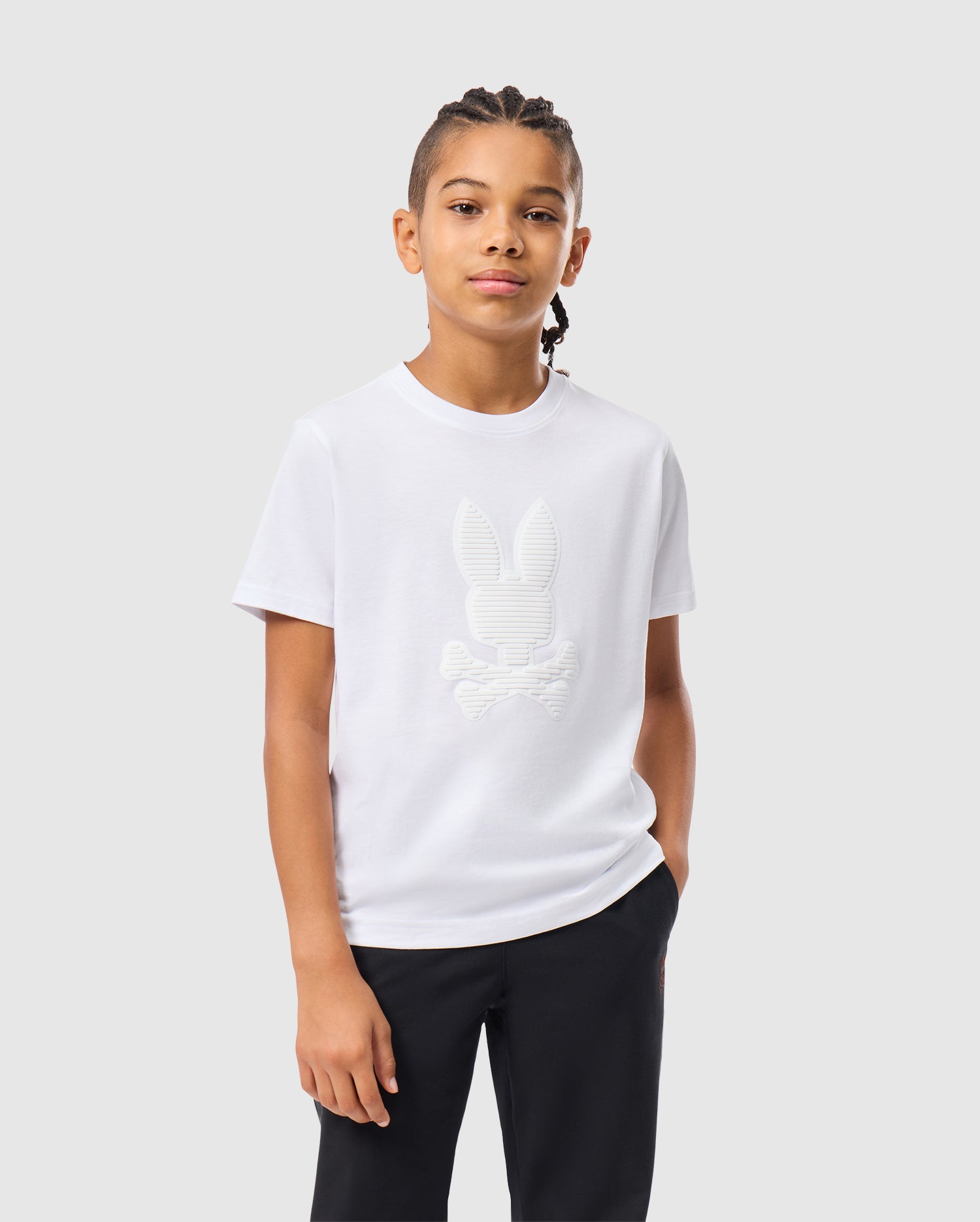 A young individual with braided hair sports a Psycho Bunny KIDS KINGSTON GRAPHIC TEE - B0U743D200 crafted from soft Pima cotton, showcasing a textured bunny and crossbones design. The white t-shirt is complemented by dark pants, creating a striking contrast against the light gray background.