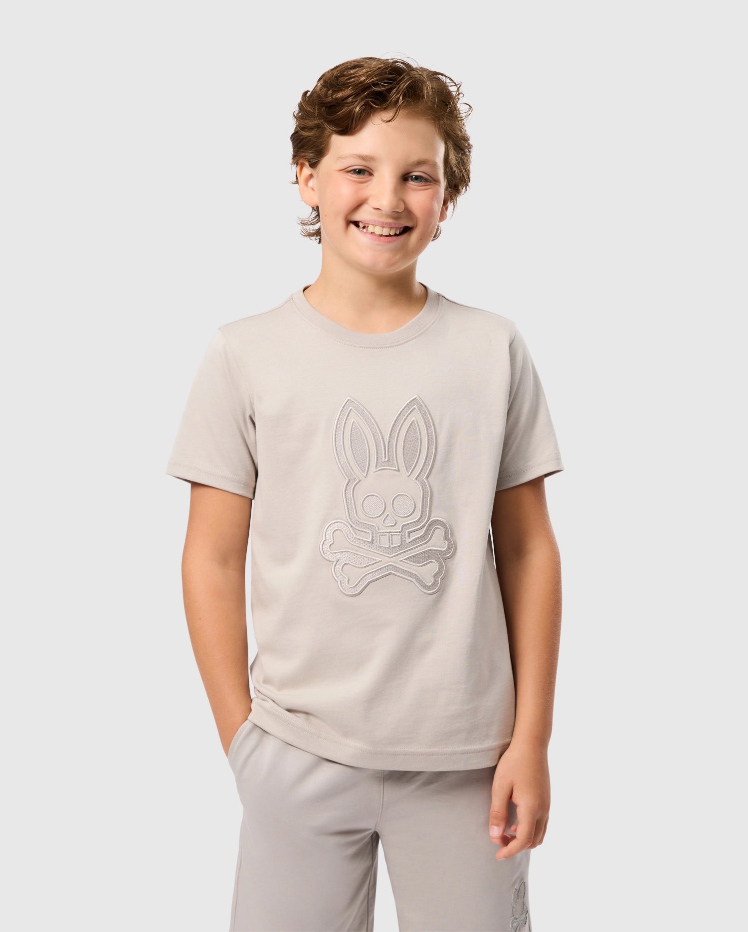 A young boy with light brown hair smiles at the camera. He is wearing a Psycho Bunny KIDS MALTON GRAPHIC TEE - B0U795D200, a light beige shirt made from soft Pima cotton, featuring a bunny skull and crossbones on the front. His left hand is in his pocket, and he stands against a plain light gray background.