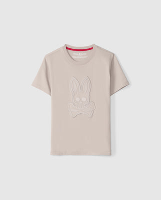 The KIDS MALTON GRAPHIC TEE - B0U795D200 from Psycho Bunny is a light beige, regular fit, short-sleeve shirt made from premium Pima cotton. It features an embossed design of a bunny's head above crossed bones and has a red inner collar with the "Psycho Bunny" brand logo inside. The tee is displayed against a plain white background.