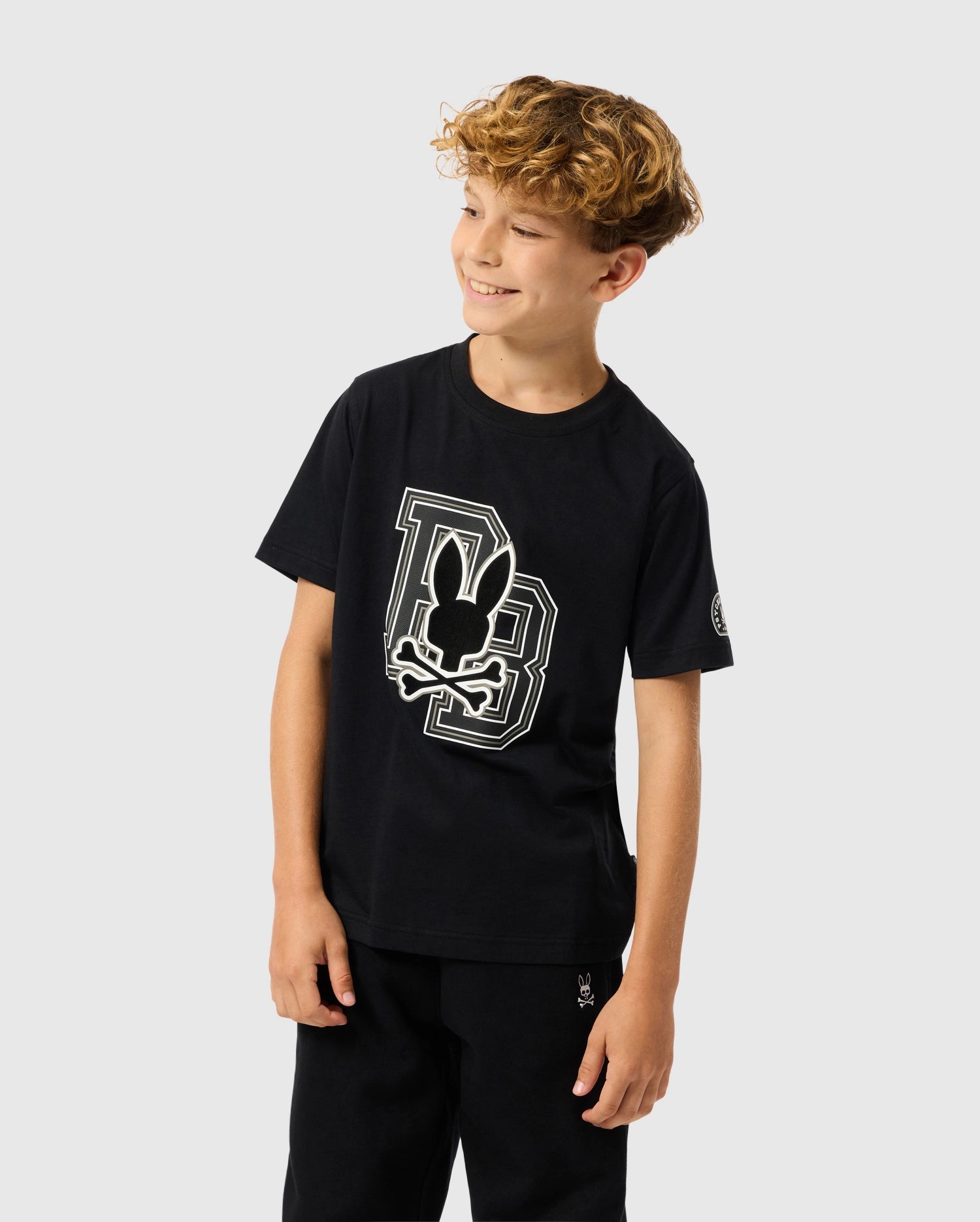 A young boy with curly hair is smiling and looking to the side, wearing a Psycho Bunny Kids Zack Graphic Tee. The black T-shirt features a stylized bunny design with the letters 