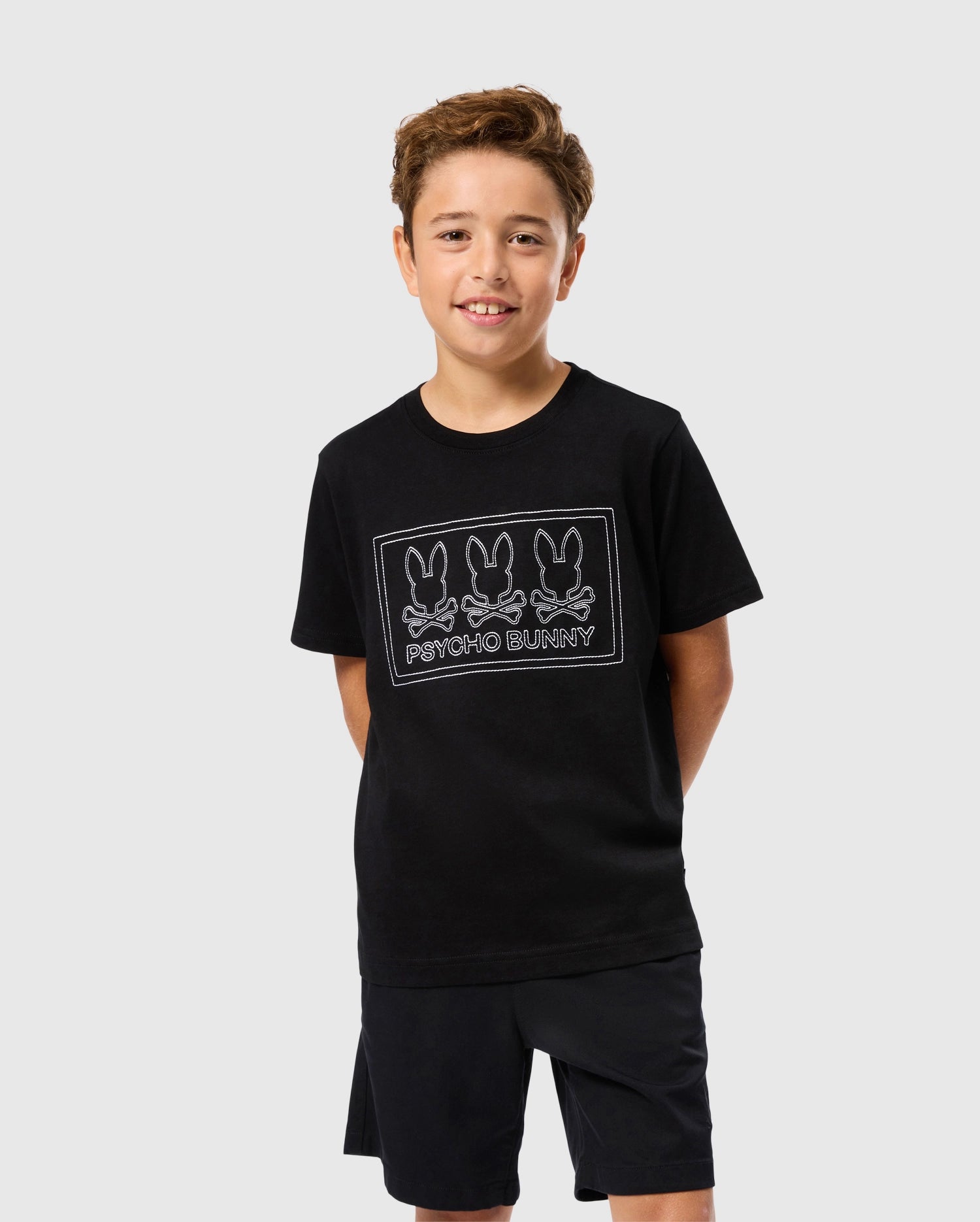 A young boy beams in a black KIDS TACOMA GRAPHIC TEE - B0U909D200 by Psycho Bunny, featuring three outlined bunny designs and embroidered details. He's paired it with matching black shorts, all made from soft Pima cotton, standing against a light gray background.