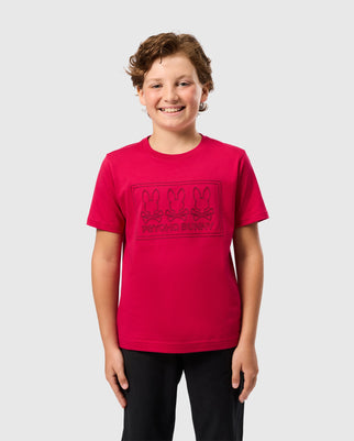 A smiling child with curly hair is wearing a bright red Psycho Bunny "Kids Tacoma Graphic Tee - B0U909D200" featuring three outlined bunny faces and the text "PSYCHO BUNNY" below them. The Pima cotton T-shirt stands out against the plain white background, with the child's hands relaxed at their sides while wearing dark pants.