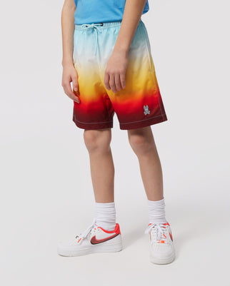 A person wearing a Psycho Bunny KIDS GUY SWIM TRUNK in shades of blue, yellow, and red stands with their hands lightly resting on their thighs. They also wear white sneakers and white socks, against a white background.