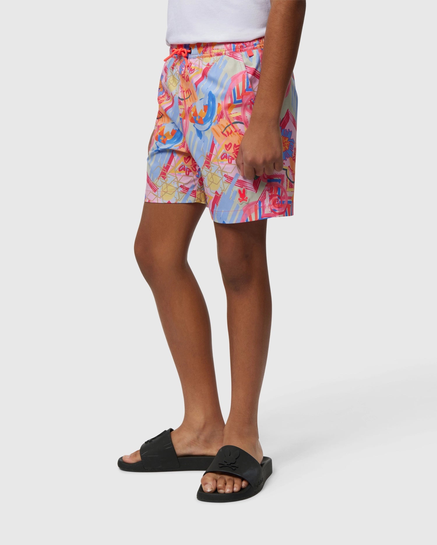 A person wearing colorful, patterned shorts and black slide sandals stands against a plain white background. The Psycho Bunny KIDS BENTON SWIM TRUNK - B0W319B2SW features a vibrant abstract design in shades of pink, orange, blue, and red.