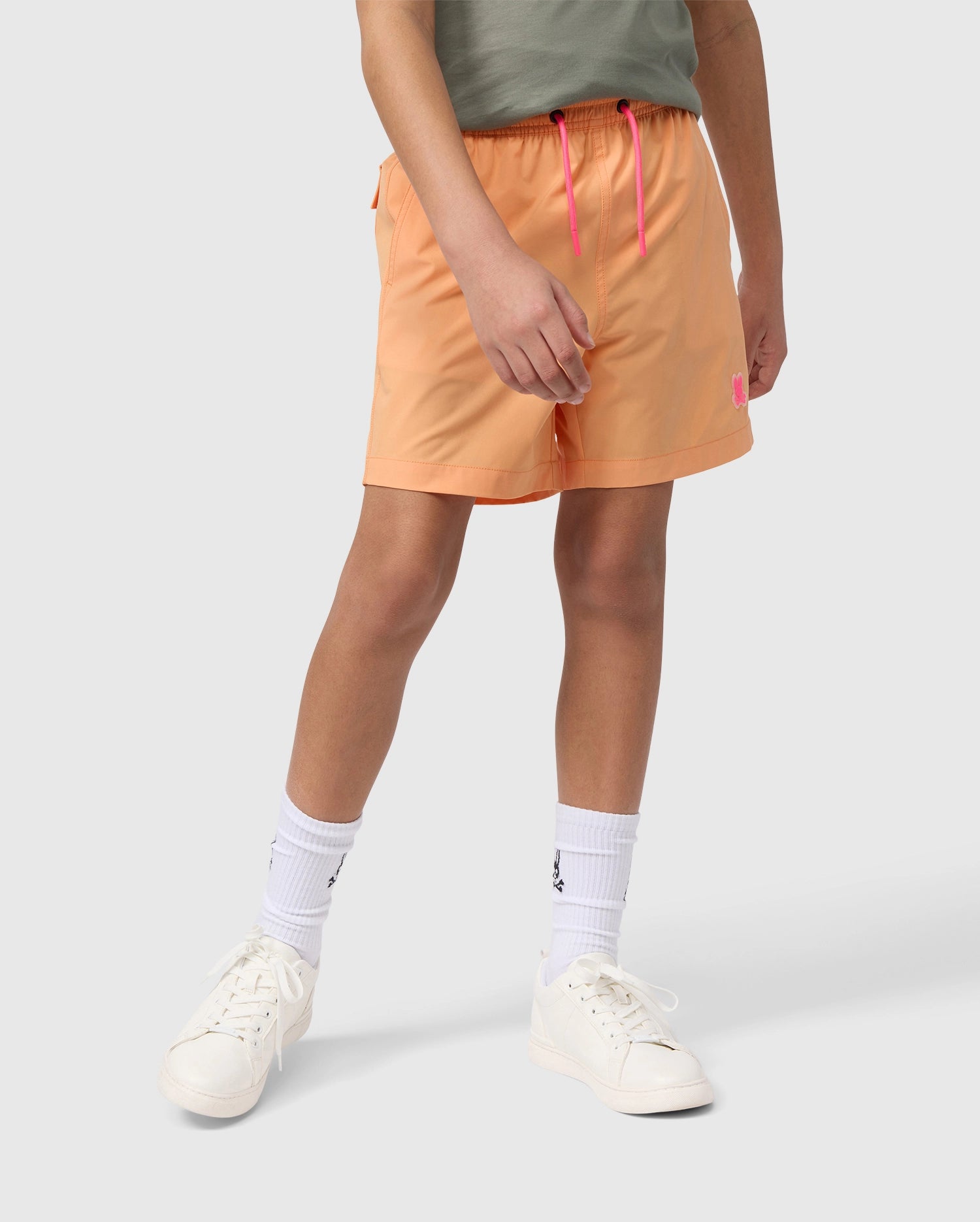 A person wearing peach-colored KIDS MALTA HYDROCHROMIC SWIM TRUNK - B0W321B2SW by Psycho Bunny with a pink drawstring and a gray t-shirt is standing with legs slightly apart. They are also wearing white socks with a small black logo and white sneakers. The background is plain light gray.