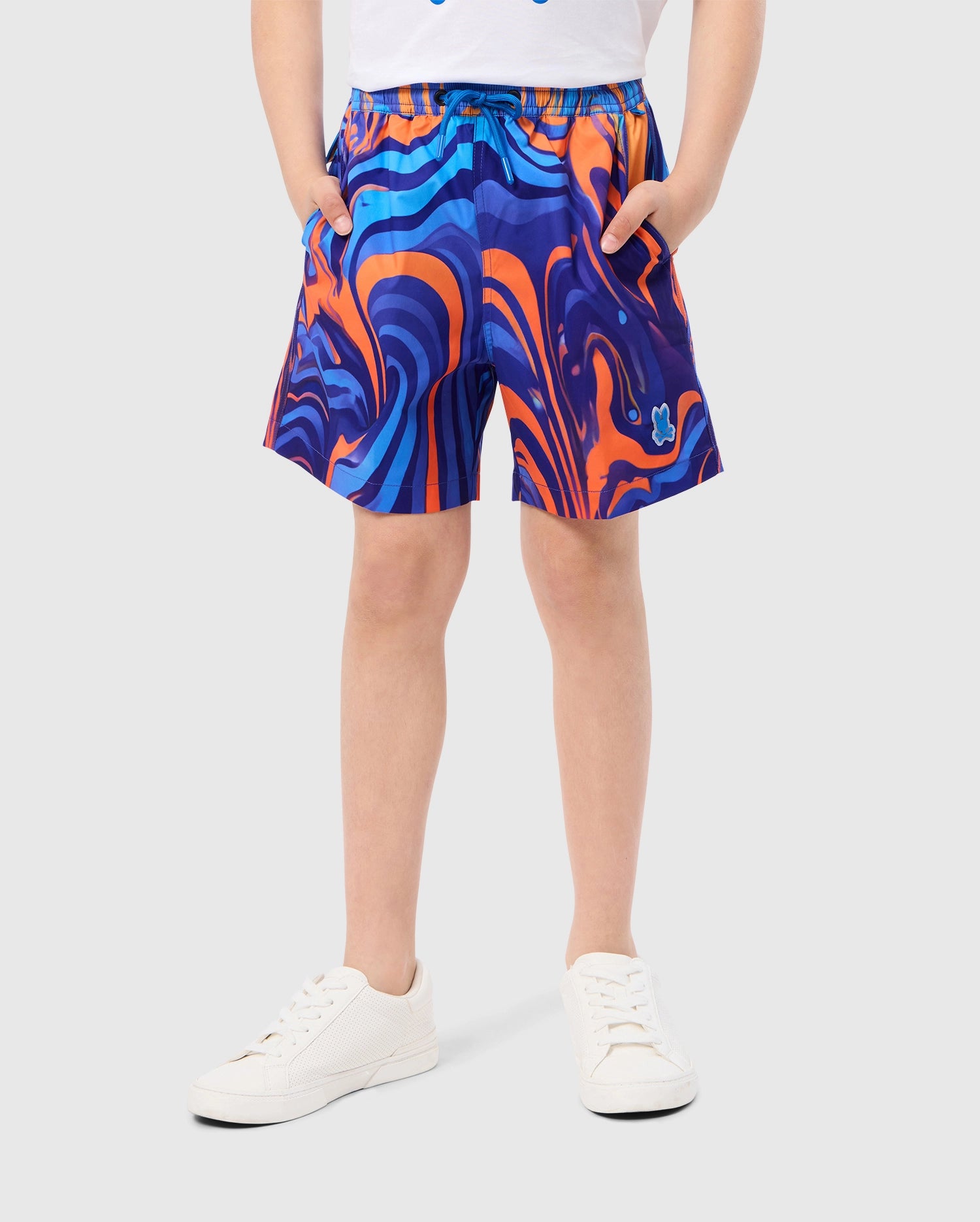 A person stands against a light grey background wearing the Psycho Bunny KIDS NEVADA ALL OVER PRINT SWIM TRUNK - B0W526C200, featuring a colorful, swirling blue and orange pattern. They have their hands in the pockets of the quick-dry fabric shorts and are also wearing a white t-shirt and white sneakers. Only the lower half of their body is visible.