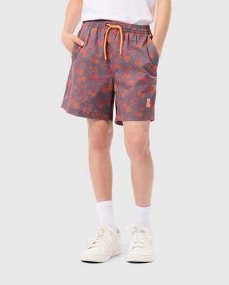 A kid wearing gray shorts with an orange bunny pattern, an orange drawstring, a white shirt, white socks, and white sneakers. The quick-dry fabric of the Psycho Bunny KIDS SHELDON ALL OVER PRINT SWIM TRUNK - B0W588C200 stands out against the plain, light-colored background.